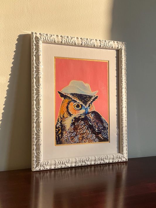 A framed original painting of a Great Horned Owl in a cowboy hat rests on a wooden surface, leaning against a blank wall, bathed in sunlight.