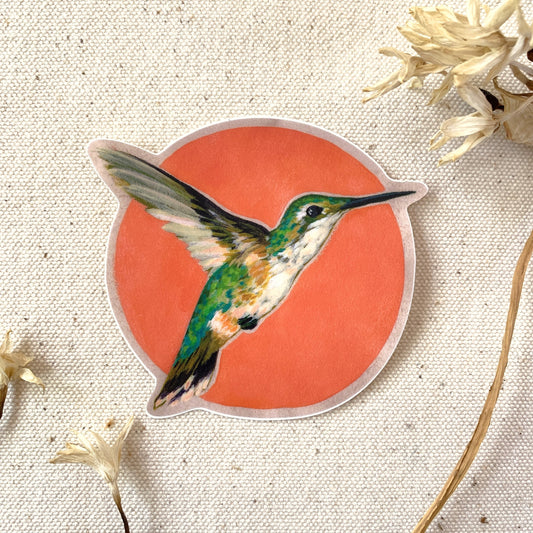 Sticker of a female hummingbird on a red circle, surrounded by dried flowers and beige fabric background.