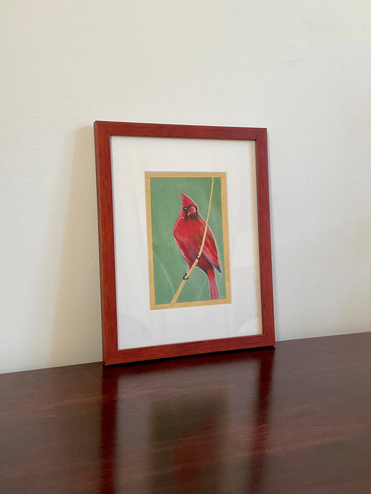 Original framed painting of a Northern Cardinal sitting on a golden branch. The painting is set on a wooden table.
