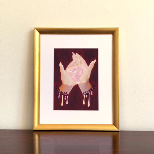 Framed artwork of hands holding a pink rose quartz crystal on a beige wall.