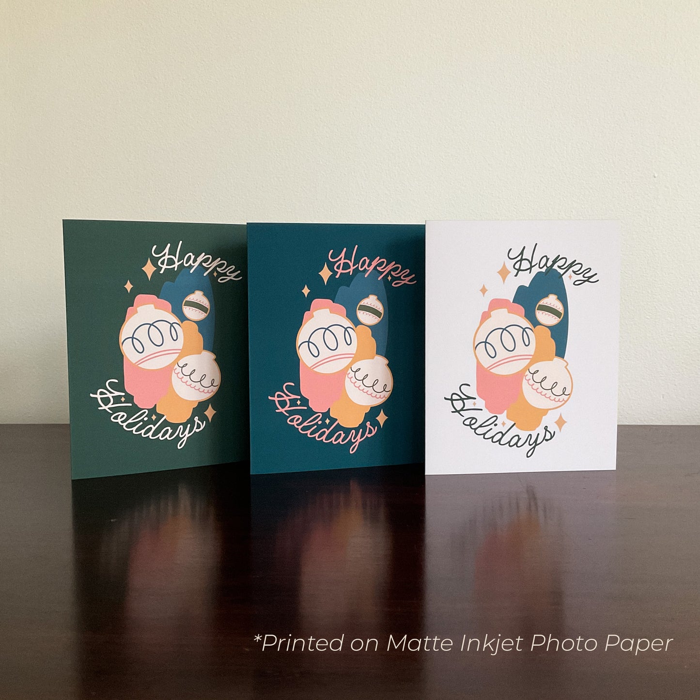 Three cards reading "Happy Holidays" with graphics of ornaments on the front, displayed on a wooden surface.