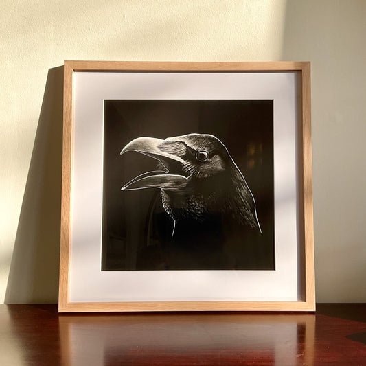 Raven Head art print framed and placed on a table, leaning against a wall. The artwork is drenched in sunlight.