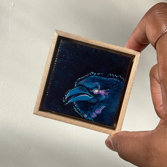 A hand holds a miniature painting of an angry blue crow, framed in a wooden float frame, slightly tilted.