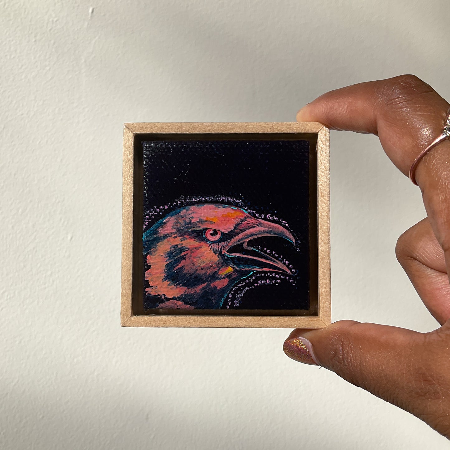 Hand holds a miniature framed artwork of a crow cawing.