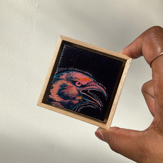 Hand holds a miniature framed painting, slightly tilted, of a crow cawing.