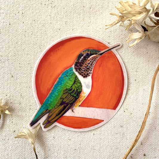 Sticker of a hummingbird on an red-orange circle, surrounded by dried flowers on a beige fabric background.