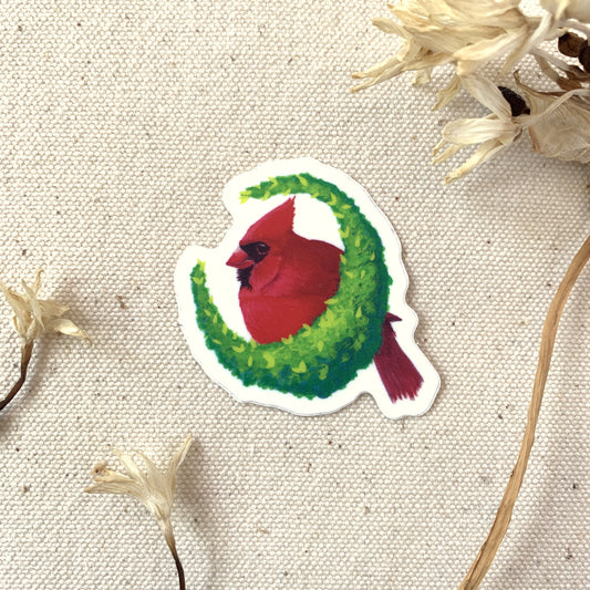 Sticker of a red cardinal bird on a green wreath surrounded by dried flowers on a textured beige background