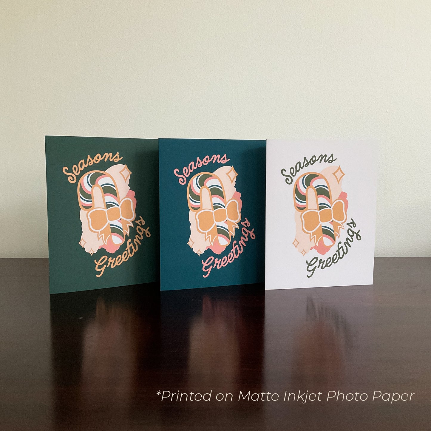Three cards reading "Seasons Greetings" with graphics of a candy cane with a bow on the front, displayed on a wooden surface.
