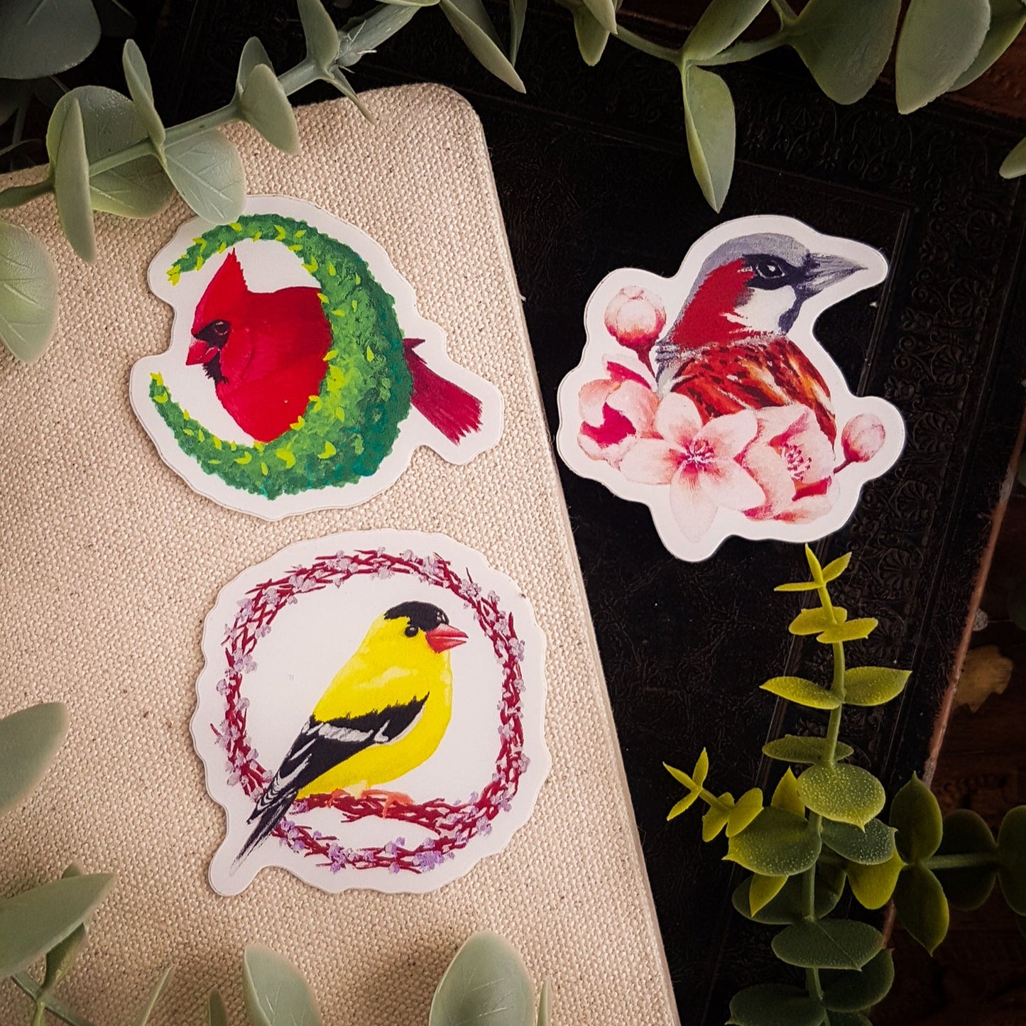 A Northern Cardinal, American Goldfinch, and House Sparrow sticker on a pile of books surrounded by greenery.