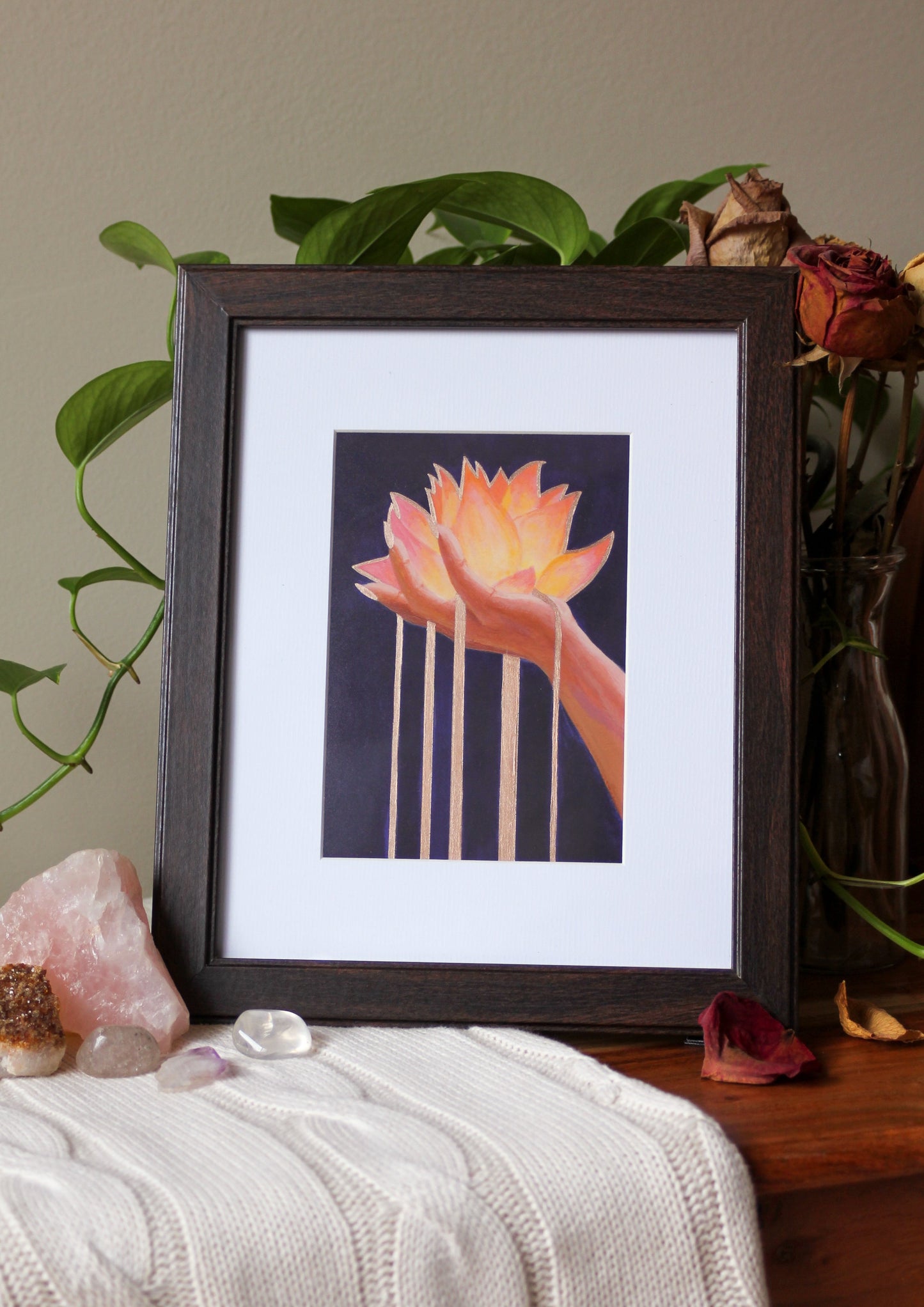 An art print of my original acrylic painting, Lotus, framed, sitting on a wooden bench, surrounded by a trailing plant, bouquet of dried roses, and crystals on a knit blanket.