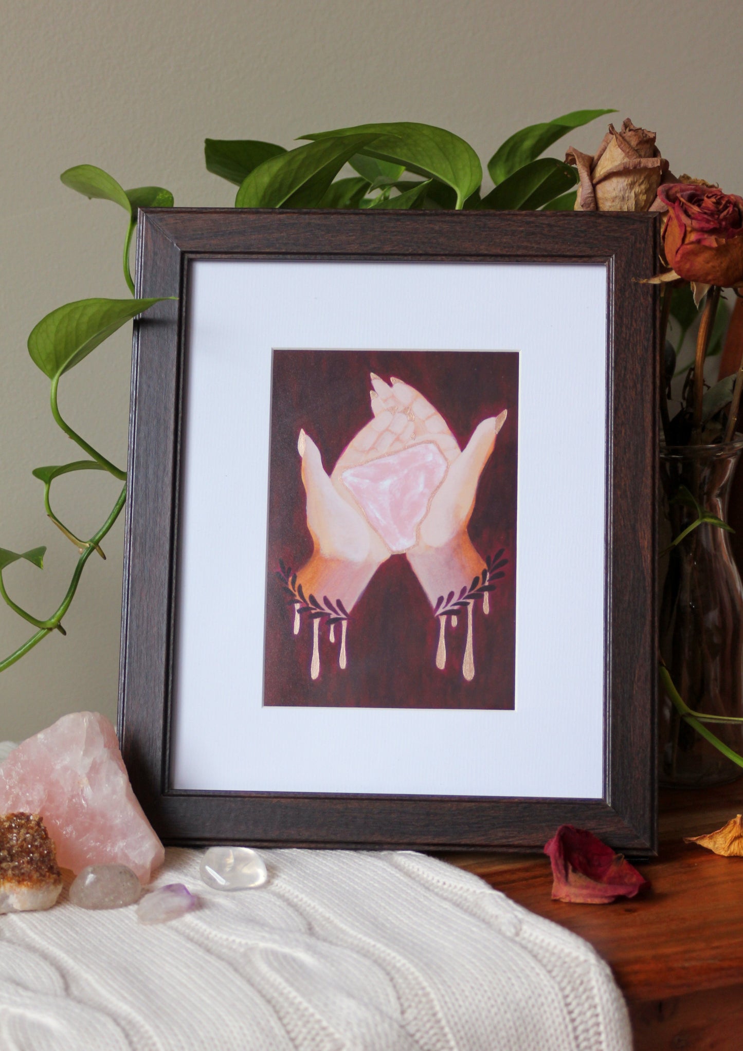 An art print of my original acrylic painting, Rose Quartz, framed, sitting on a wooden bench, surrounded by a trailing plant, bouquet of dried roses, and crystals on a knit blanket.