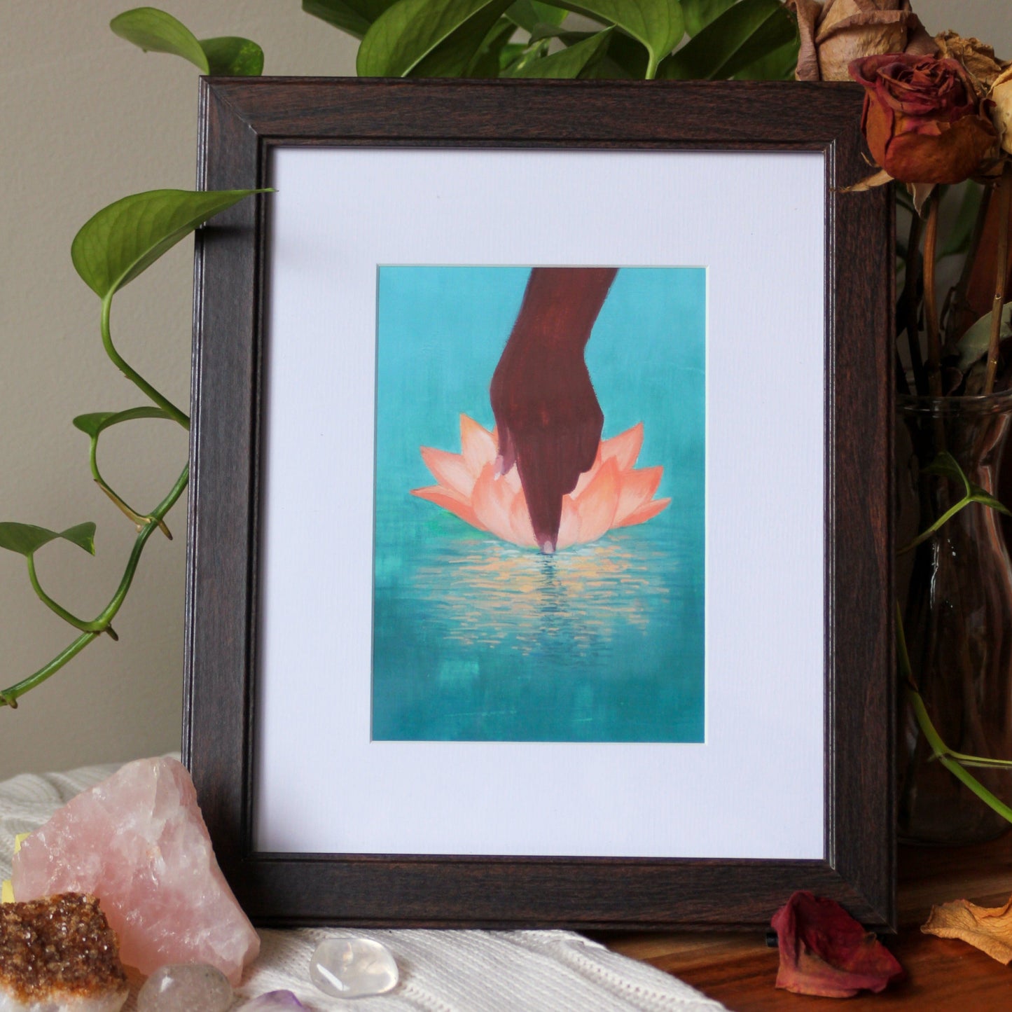 An art print of my original gouache painting, Bloom, framed, sitting on a wooden bench, surrounded by a trailing plant, bouquet of dried roses, and crystals on a knit blanket.