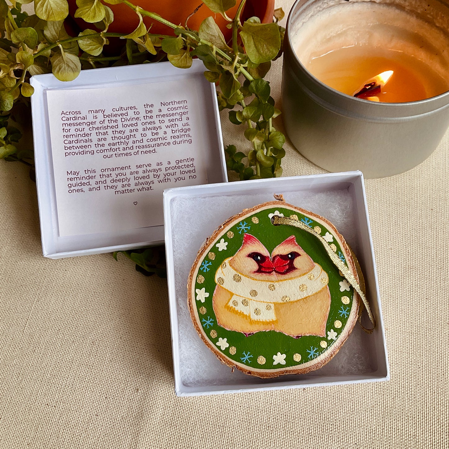 Decorative, hand-painted ornament in open box with notecard on a beige cloth with a candle.