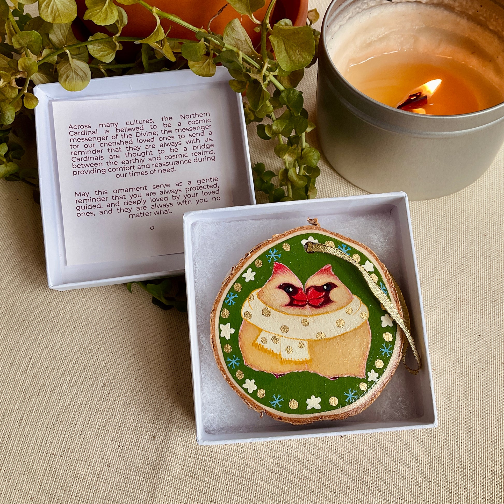 Decorative, hand-painted ornament in open box with notecard on a beige cloth with a candle.