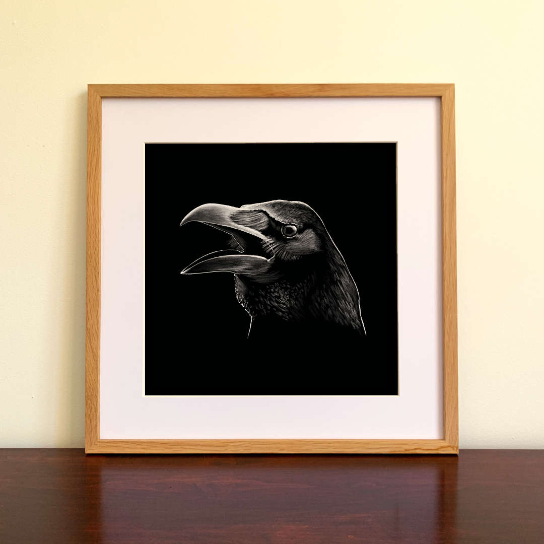 Framed black and white drawing of a raven on a wooden surface with a beige wall background.