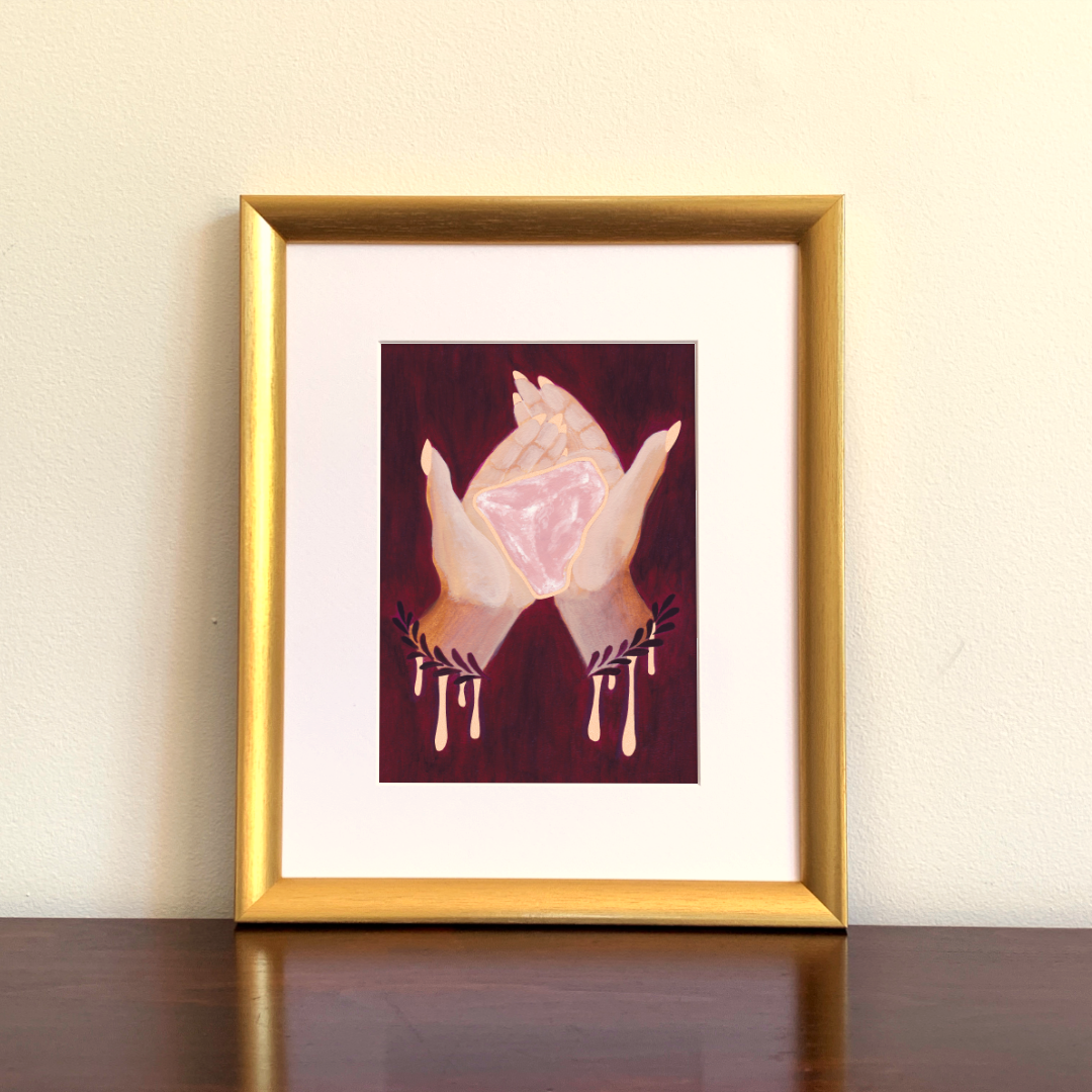 Framed artwork of hands holding a pink rose quartz crystal on a beige wall.
