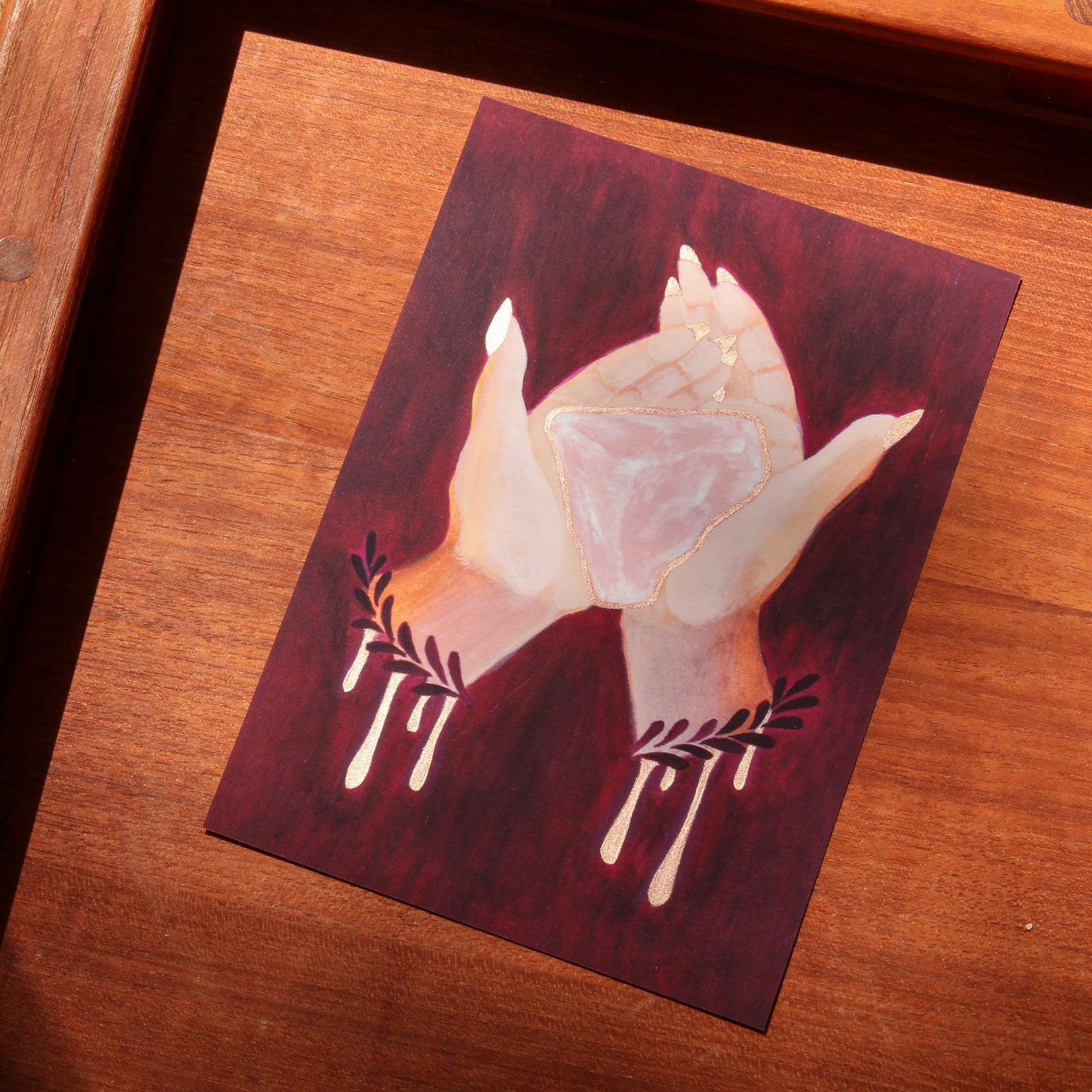 Art print of hands holding a pink rose quartz crystal on a wood surface. 