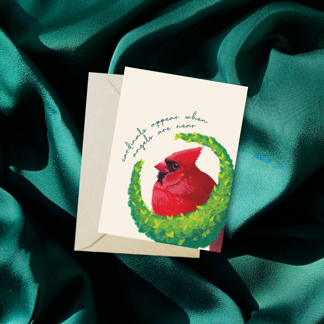 Miniature greeting card and envelope with red cardinal bird on a green wreath against teal fabric.