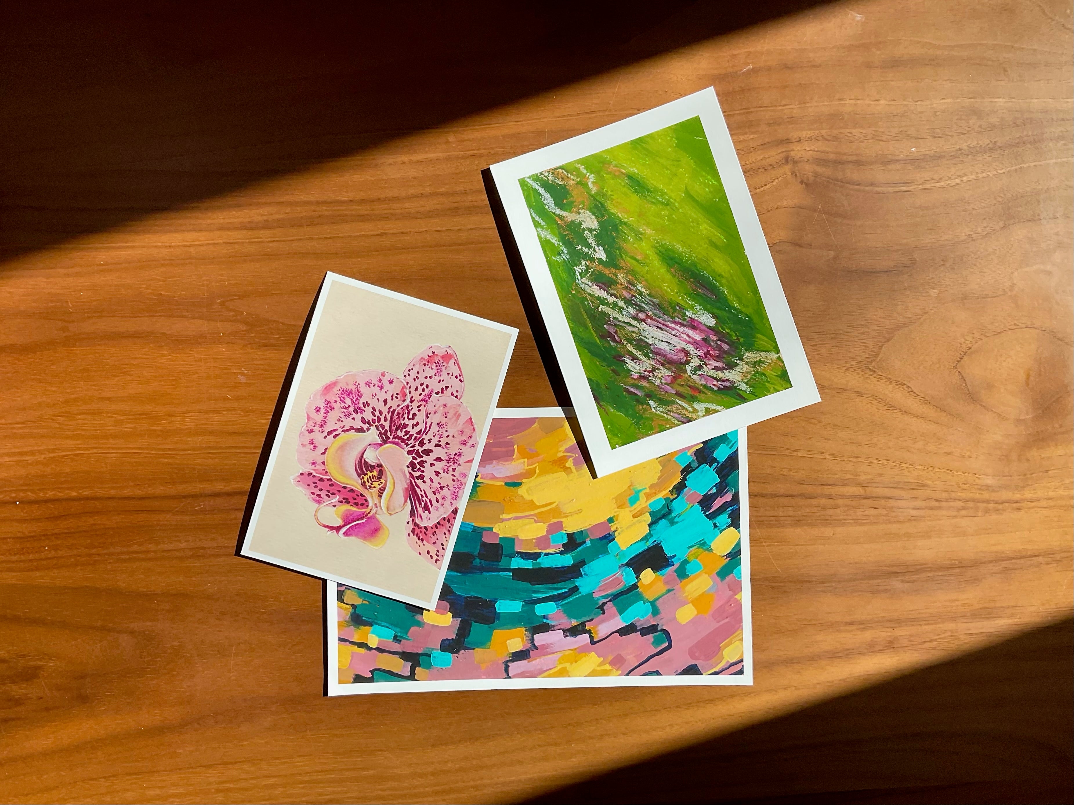 Three art prints on a wooden surface, sunlight washing over them.