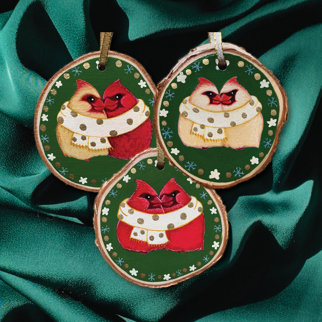 Three wood slice ornaments with Northern Cardinals wrapped in a cozy scarf painted on them on top of a silk background.